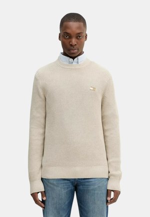 Suéter de punto beige con cuello y puños acanalados, que presenta un pequeño detalle de logo en el pecho. Lleva encima una camisa de botones azul claro.