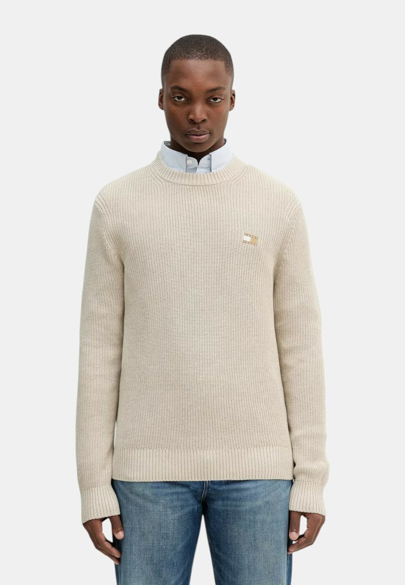 Maglioncino beige a maglia con colletto e polsini a costine, con un piccolo logo sul petto. Indossato sopra una camicia azzurro chiaro con bottoni.