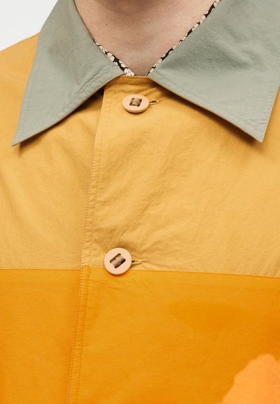 Πουκάμισο με κουμπιά και πράσινο γιακά, με σχέδιο πατς εργασίας που περιλαμβάνει μουσταρδί και πορτοκαλί χρωματικά μπλοκ.