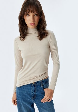 Haut à manches longues beige à col roulé décontracté, fabriqué en tissu doux et extensible ; associé à un jean taille haute bleu clair. Design lisse et ajusté.