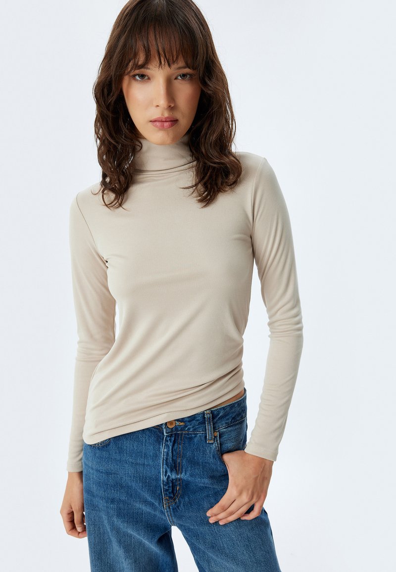 Beige mock turtleneck pitkähihaine paita, joka on valmistettu pehmeästä ja venyvästä kankaasta; yhdistetty vaaleansinisiin korkeavyötäröisiin farkkuihin. Suora, istuva muotoilu.