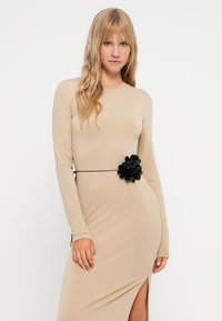 Beige langermet kjole med sideslipp, pyntet med en svart blomst beltedetalj, laget av glatt stoff med en tettsittende silhuett.