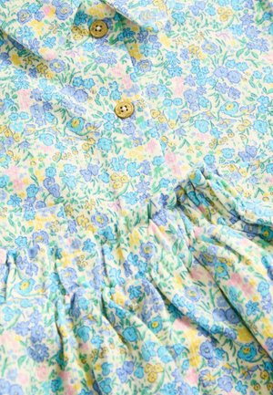 Tissu à motif floral dans des couleurs pastel, présentant de petites fleurs bleues sur un fond jaune clair. Deux boutons marron ornent le col.