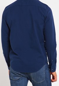Marineblauwe overhemd met lange mouwen en een button-down kraag, eenvoudig ontwerp en lichte glans. Heeft knoopmanchetten en een afgeronde onderkant.