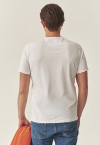 Fehér rövid ujjú póló kerek nyakkivágással, sima textúrával. Kék farmerrel viselve, narancssárga dzsekivel az alkarra terítve.