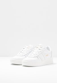 Sneakers bianchi con tomaia in pelle liscia, caratterizzati da sottili strisce grigie e una suola bianca rialzata. Logo Gola in arancione chiaro sul lato.