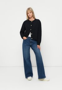 Svart kort cardigan med teksturert stoff, med gullknapper. Kombinert med mid-blue vide jeans og hvite sneakers for en uformell stil.