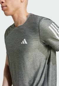 Camiseta deportiva de color gris oscuro con una tela texturizada. Presenta acentos en blanco con tres rayas en los hombros y un logo sutil en el pecho.