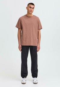 Braunes Baumwoll-T-Shirt mit kurzen Ärmeln und rundem Halsausschnitt; kombiniert mit schwarzen, schmalen Jogginghosen. Die Sneakers haben weiße und blaue Akzente.