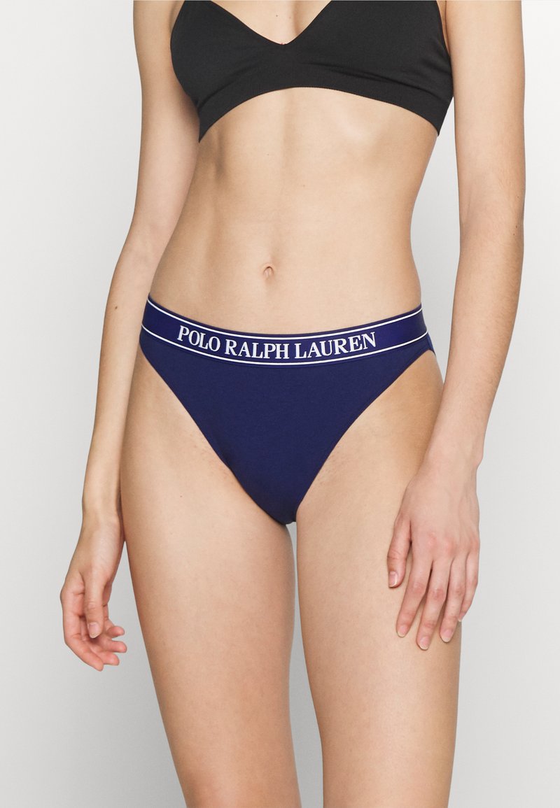 Tumedas sinine puuvillane string, millel on valge logo vöö. Disain sisaldab siledat tekstuuri ja mõõdukat katteala. Koos musta braletiga.