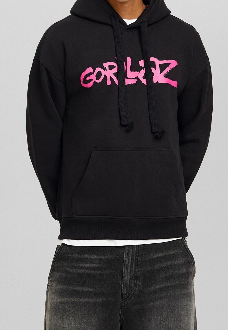 Svart hoodie med rosa "GORLZ"-text, snörning i huvan, framficka och ribbad nederdel. Materialet verkar mjukt och texturerat.