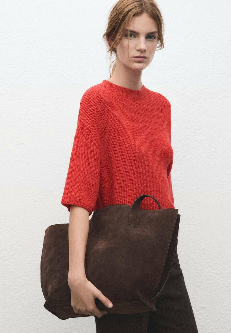 Sac cabas en daim marron avec une large ouverture et une anse courte, associé à un pull en maille rouge uni. Le mannequin porte un pantalon sombre ajusté.