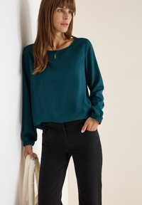 Türkisfarbige Bluse mit rundem Ausschnitt und Schlüsselelement, ausgestattet mit langen Ärmeln und elastischen Bündchen, kombiniert mit schwarzen Slim-Fit-Jeans.