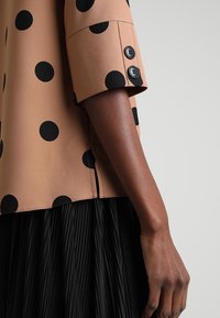 En arm med mörk hudton som bär en tanblus med stora svarta prickar och två svarta knappar vid manschetten, tillsammans med en svart, plisserad kjol.