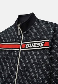 Giacca bomber nera con un motivo a logo ripetuto, striscia rossa con il testo 'GUESS', chiusura con zip e colletto a costine.