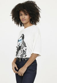 Weißes, übergroßes T-Shirt mit schwarzem und blauem Grafikdesign eines Gesichts, umgeben von Blumen. Dazu werden dunkle Jeans und ein besetzter schwarzer Gürtel getragen.