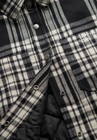 Sort-hvid ternet skjorte med fleece-struktur, der har metaltrykknapper og quiltet sort foret. Inkluderer to forlommer.