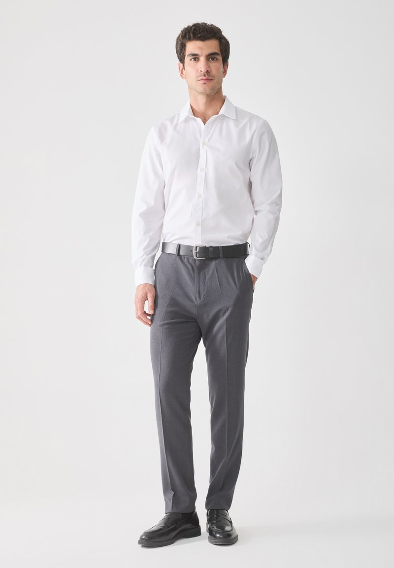 Witte overhemd met knopen, grijze broek, zwarte leren riem en zwarte schoenen. Minimalistisch ontwerp, gladde stof, aansluitende pasvorm.