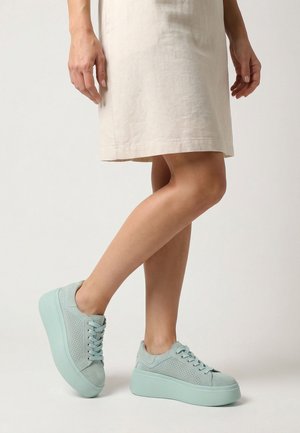 Legs in beige knee-length skirt wearing light blue platform sneakers, one foot slightly raised on a light background.