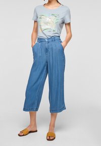 Woman wearing light blue tropical print t-shirt, high-waist wide-leg cropped denim pants, and tan woven slide sandals, hands in pockets.