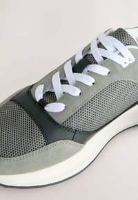 Close-up af en grå og sort sneaker med hvide snørebånd, med mesh og ruskindstekstur og en hvid gummisål.