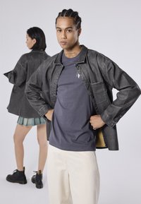 Giacca di jeans nera con vestibilità rilassata, t-shirt grigia, pantaloni bianchi e stivali neri con dettagli gialli; il modello posa con sicurezza.