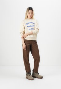 Hanorac crem cu text albastru, pantaloni maronii și cizme gri. Hanoracul are manșete strâmte; cizmele au șireturi și tălpi texturate.