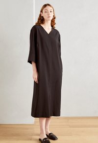 Robe noire ample avec un décolleté en V et des manches trois-quarts. Tissu lisse avec une coupe longue et droite jusqu'aux chevilles.