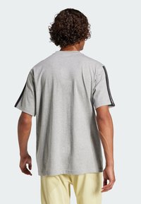 Šedé tričko s krátkym rukávom, ktoré má čierne bočné pruhy a voľný strih, vyrobené z jemnej bavlny. Pohľad zozadu odhaľuje hladkú textúru a jednoduchý dizajn.