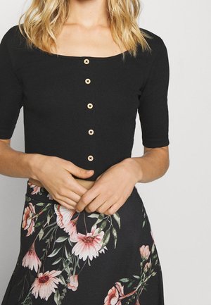 Blonde Frau, die ein schwarzes kurzes Oberteil mit Knöpfen und einen schwarzen Rock mit großen pinken und grünen Blumenmustern trägt.