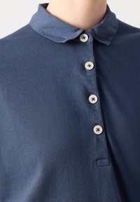 Chemise col bleu marine avec texture côtelée, dotée d'une patte de boutonnage à six boutons et de boutons beiges. Design minimaliste avec une poche sur le côté gauche.