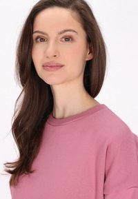 Lyserød sweatshirt med rund hals, afslappet pasform og blød tekstur. Modellen har langt brunt hår og minimal makeup. Baggrunden er helt hvid.