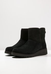 Botines de ante negro con puntera redondeada y suela baja de cuña. Textura suave, costuras mínimas y un tirador en la parte posterior.