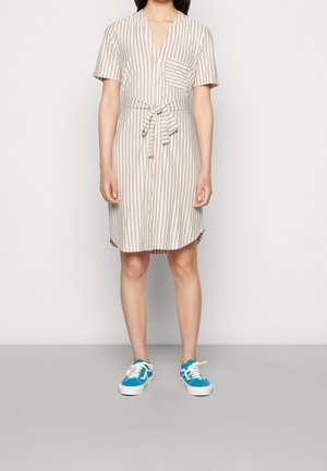 Personne portant une robe boutonnée beige et blanche à rayures verticales, manches courtes, ceinture à la taille et baskets bleues avec lacets blancs.
