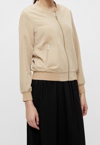 Veste aviateur beige avec un col rond, une fermeture éclair frontale et des poignets élastiques. Tissu lisse avec une taille ajustée, associée à une jupe noire plissée.