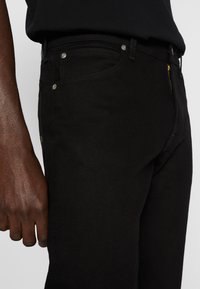 Tume nahk käsi, mis resting mustade teksade kõrval, millel on nähtavad hõbedased nupud ja osaliselt nähtav kuldne tõmblukk, valge tausta ees.