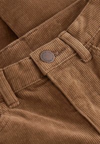 Tissu en velours côtelé marron avec une texture à nervures. Présente un bouton en métal avec un texte gravé et des passants de ceinture. Gros plan de la zone de taille.