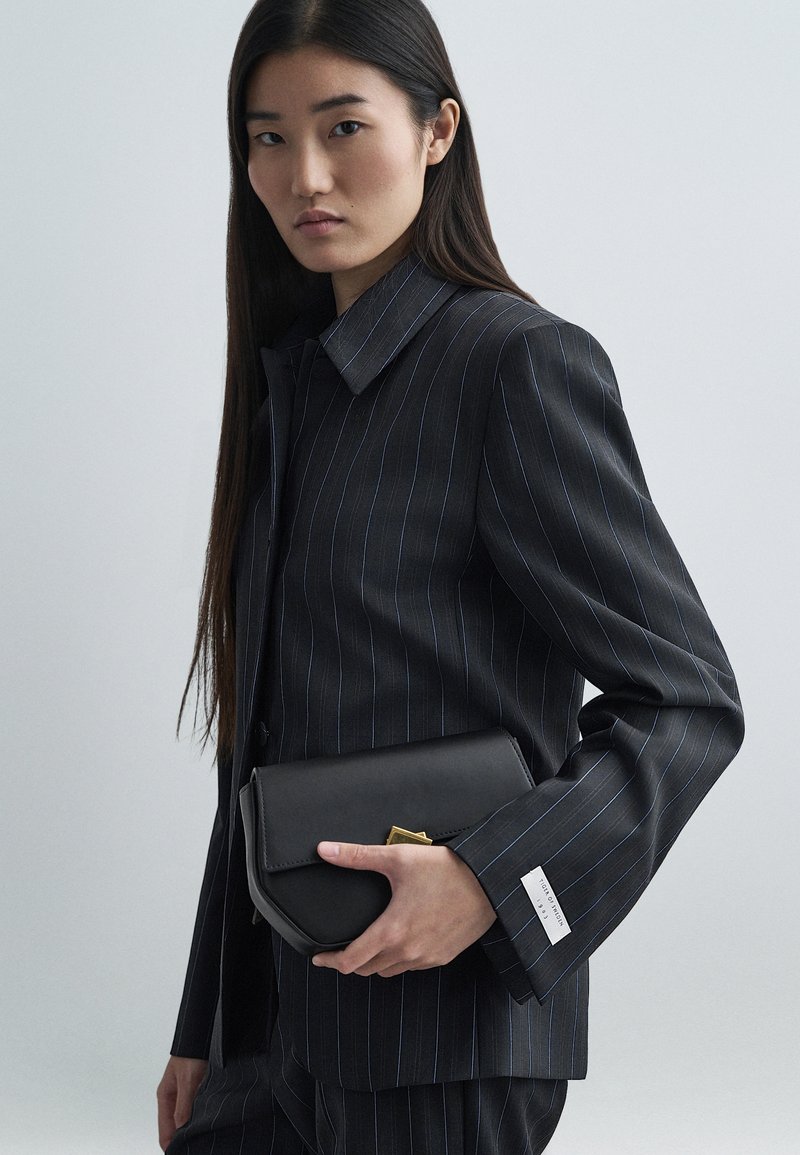 Zwarte leren clutch met een geometrisch gouden accent, gecombineerd met een donker gestreept pak met blauwe lijnen en een overhemdkraag.