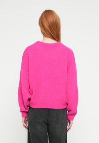 Spilgti rozā adīts džemperis ar vaļīgu piegriezumu, ribots apakšmala un apaļa kakla izgriezums, kopā ar tumši pelēkām atslābināta piegriezuma biksēm.