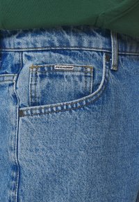 Vaaleansiniset farkut, joissa on kulunut pinta, ja takataskussa pieni mustavalkoinen PEGADOR-tunnus. Näkyvissä on kontrastivärinen ompelu.