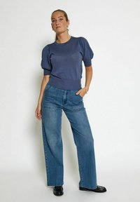 Jūrų pajūrio pūkuotų rankovių trikotažo viršus su šerpetoto apačia, derinamas su aukštu juosmeniu, plačiomis mėlynomis džinsomis. Aprangą užbaigia juodi batai.