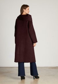 Lang, donker bordeauxrood jasje van zacht materiaal, met een brede kraag en losse mouwen, gecombineerd met uitlopende blauwe jeans en hoge hakken.