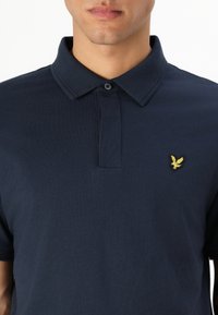 Jūros mėlynos spalvos medvilninė polo marškinėliai su sagomis apykakle ir mažu geltonu erelio logotipu kairėje krūtinės pusėje. Lygus paviršius, trumpomis rankovėmis.