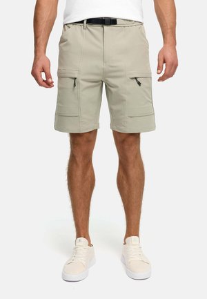 Mężczyzna w beżowych spodniach typu cargo z zamkami błyskawicznymi w bocznych kieszeniach, czarnym paskiem i białymi tenisówkami.