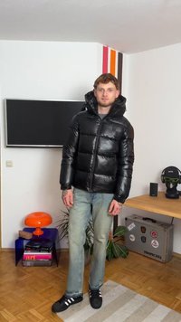 Casaco puffer preto com capuz, apresentando textura acolchoada e fecho com zíper. Combinado com jeans azul claro e sapatos pretos com riscas brancas.