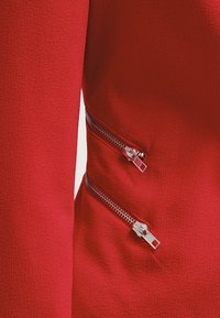 Chaqueta ajustada roja con tejido texturizado y dos cremalleras plateadas horizontales a los lados, añadiendo un detalle funcional al diseño.