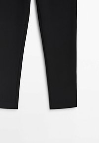 Partie inférieure d'un pantalon noir ajusté à jambes droites sur fond blanc, montrant la texture du tissu et les détails des coutures.