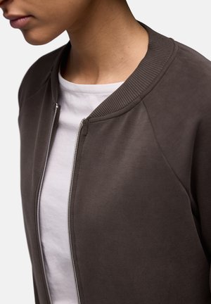 Giacca bomber marrone con collo a coste, chiusura con zip e texture liscia, indossata sopra una maglietta bianca. Focus sulla vestibilità rilassata e sulla semplicità.