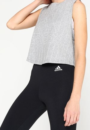 Femme portant un haut gris sans manches Adidas et des leggings noirs Adidas, posant avec une main près du cou et l'autre sur le côté.