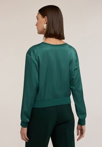 Maglione in satin teal con vestibilità rilassata, polsini e vita a costine. Presenta un ampio scollo e una texture morbida. Indossato con pantaloni abbinati.